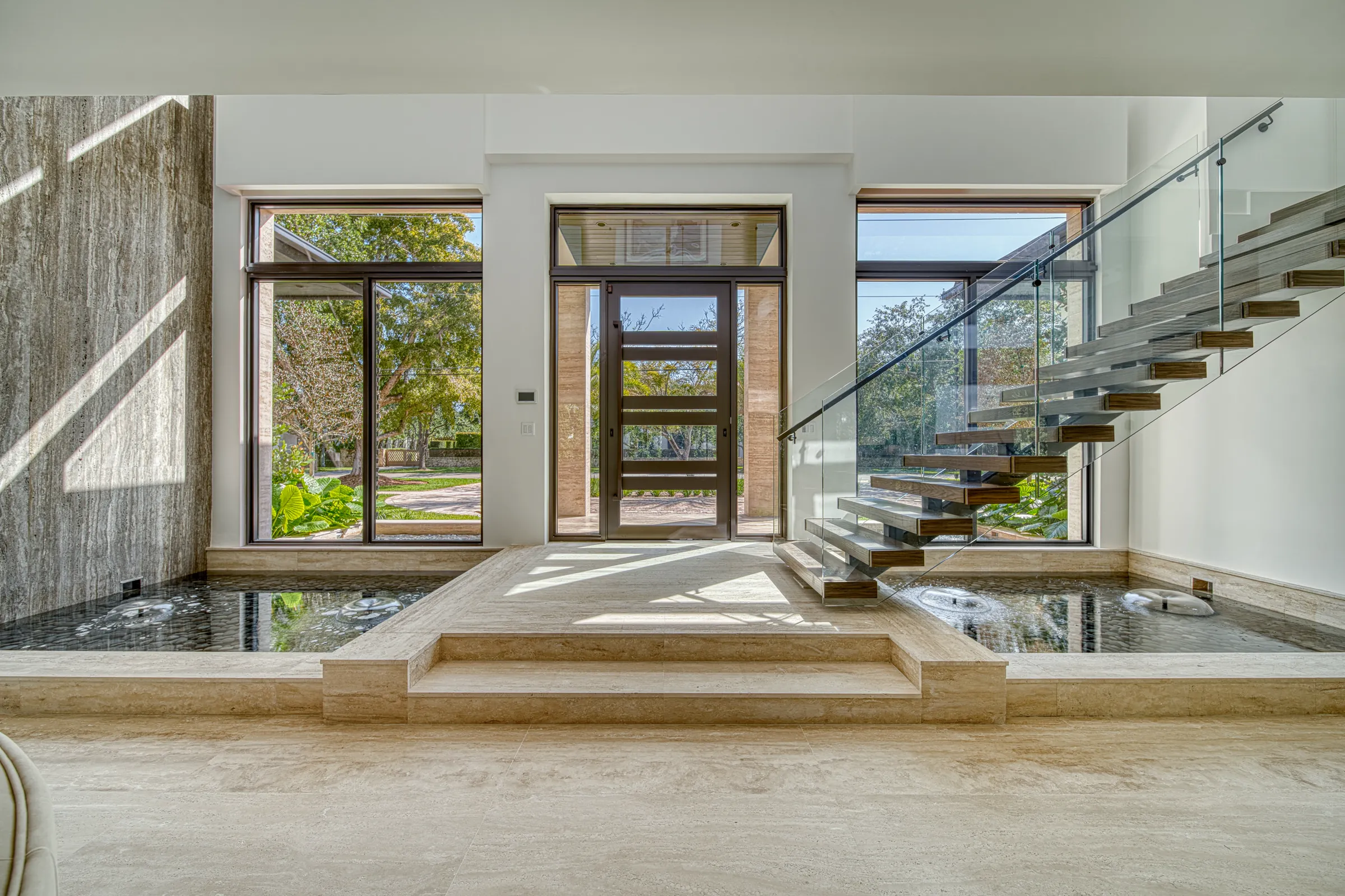 Sunlit travertine entry with a floating glass staircase and reflecting pools
